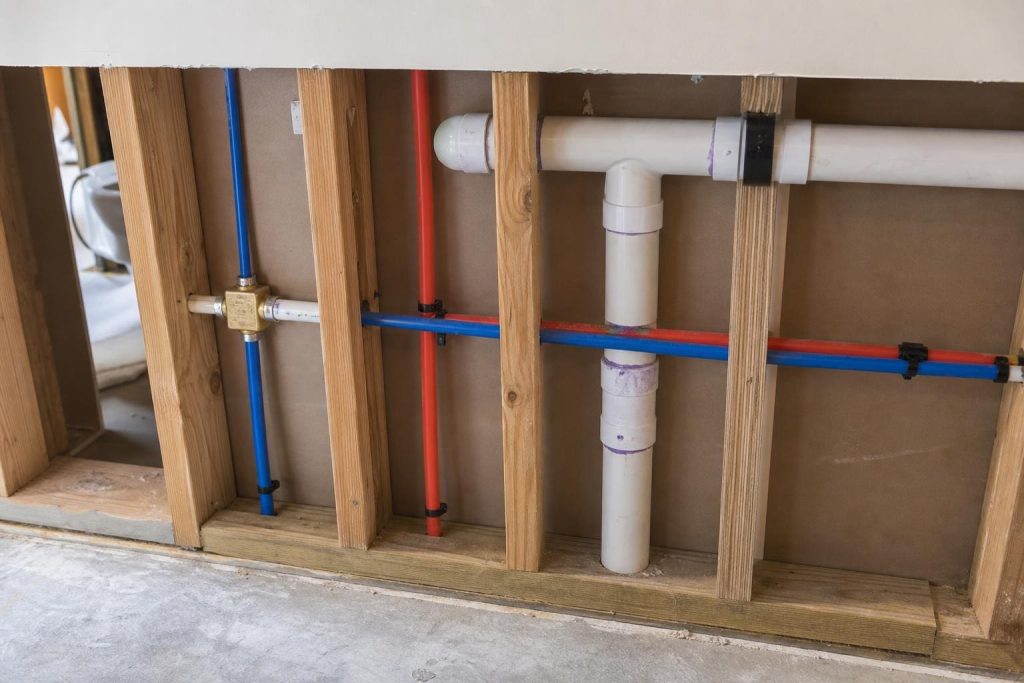 Exposed wall framing showing a network of red and blue PEX water lines alongside white PVC drain pipes and a brass mixing valve.