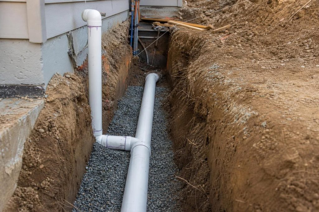 White PVC pipes installed in a deep dirt trench with gravel bedding alongside a residential building foundation.